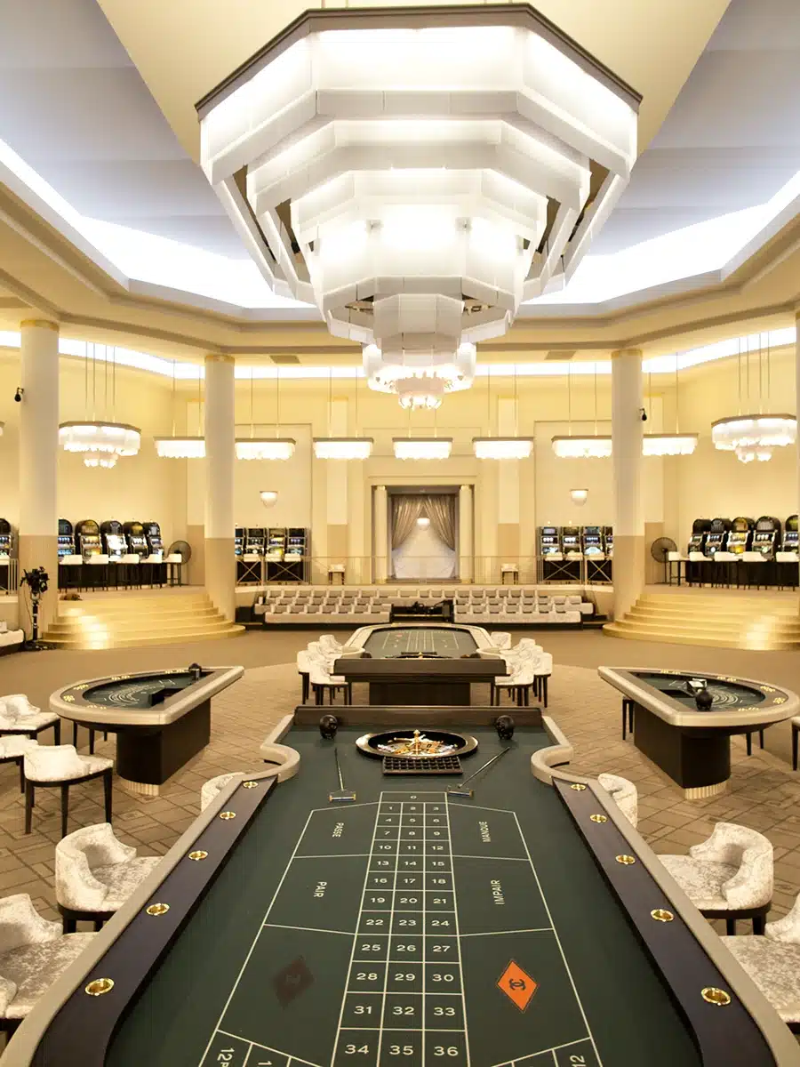 Cette image montre un somptueux décor de salle de casino art déco, recréé en studio. On y voit de grandes tables de jeu ovales au centre, entourées de fauteuils élégants. L’espace est baigné d’une lumière dorée provenant de multiples lustres géométriques suspendus au plafond, eux-mêmes intégrés à une architecture aux lignes arrondies et raffinées. Des gradins disposés tout autour accueillent les spectateurs, tandis que des colonnes blanches rythment l’ensemble. L’atmosphère évoque le luxe feutré d’un grand palace des années 1930, parfaitement mis en scène.