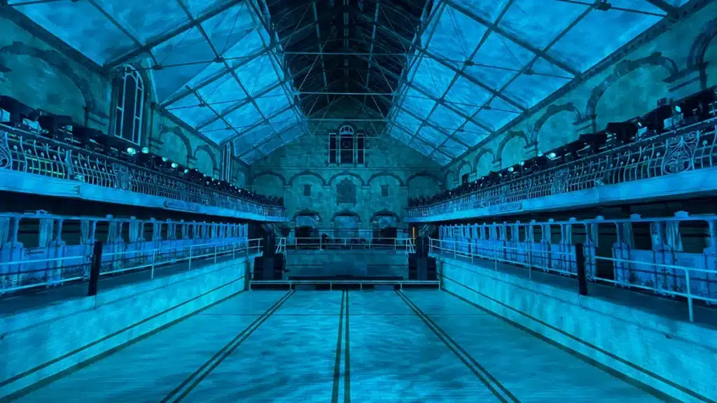 Photo représentant un décor de piscine vide. Projection d'une image sur un écran. Coussins et tapis ancien au sol.