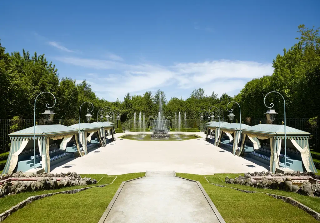 Un jardin à la française avec une grande fontaine centrale, entouré de pavillons élégants aux toits bleu-vert, décorés de rideaux crème et de coussins bleus. L’ensemble évoque un espace extérieur raffiné, conçu pour accueillir des invités dans une ambiance aristocratique.