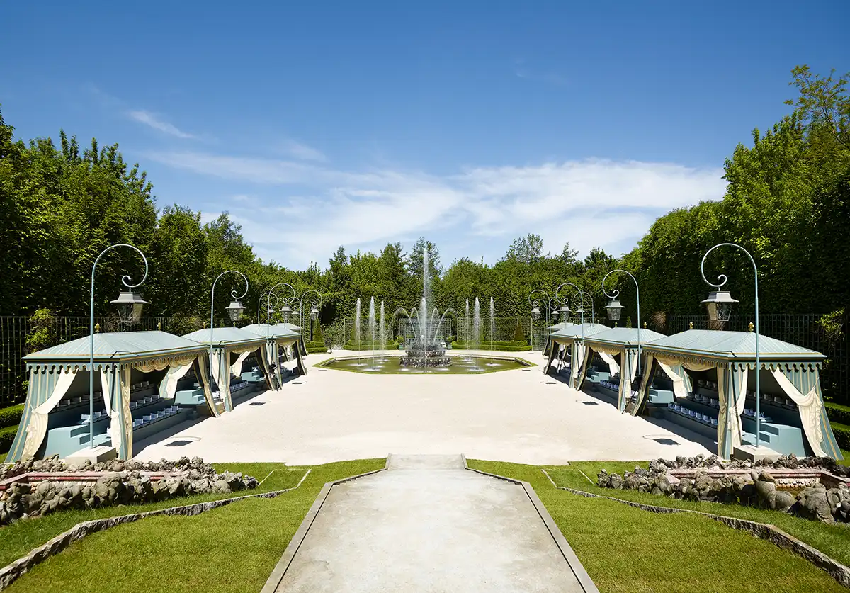 Un jardin à la française avec une grande fontaine centrale, entouré de pavillons élégants aux toits bleu-vert, décorés de rideaux crème et de coussins bleus. L’ensemble évoque un espace extérieur raffiné, conçu pour accueillir des invités dans une ambiance aristocratique.