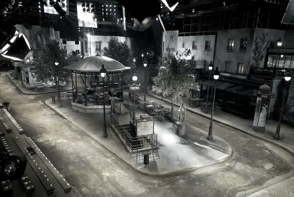 La scène représente la reconstitution minutieuse d’une rue parisienne rétro, entièrement construite en décor de studio. On y voit un kiosque à musique au centre, entouré de lampadaires haussmanniens, d’arbres, de terrasses de cafés et d’un entrée de métro ancienne avec sa grille caractéristique. Les façades des immeubles, patinées et légèrement usées, évoquent un Paris du début du XXᵉ siècle. L’atmosphère est volontairement cinématographique, avec un éclairage contrasté qui accentue les textures du sol et des murs. Des gradins disposés sur le côté indiquent qu’il s’agit d’un décor de défilé, pensé pour accueillir un public.