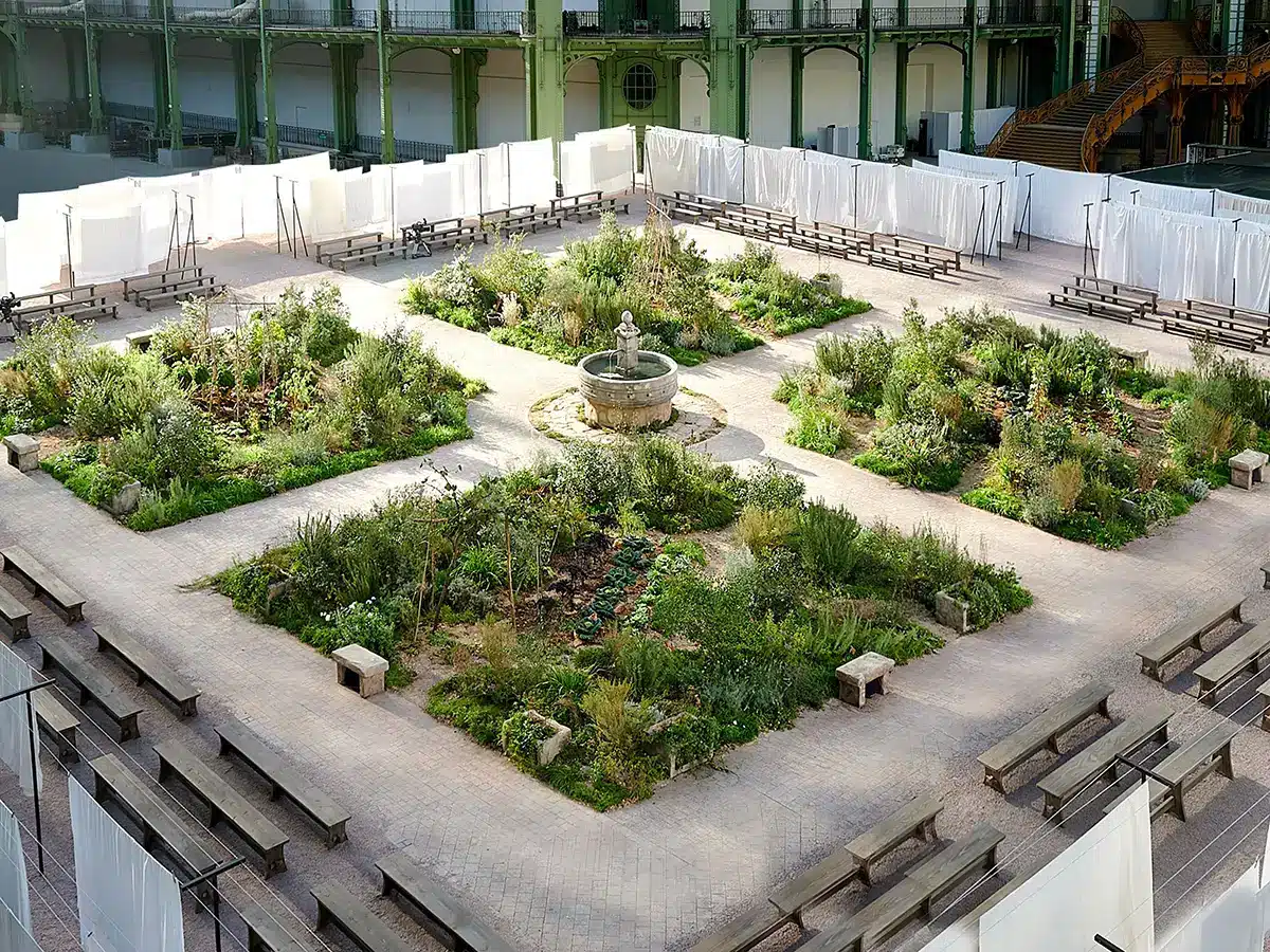 Cette image montre une vaste scénographie installée sous la verrière du Grand Palais, transformé ici en jardin à ciel couvert. L’espace est aménagé comme un cloître végétal : quatre grands massifs de plantes sauvages et méditerranéennes forment une croix symétrique, organisés autour d’une fontaine en pierre ancienne placée en son centre. Les parterres, denses et luxuriants, mélangent buissons, herbes aromatiques, fleurs champêtres et arbustes, donnant l’impression d’un jardin rural ou monastique, volontairement laissé à l’état naturel. Les chemins pavés qui séparent les massifs créent des allées où les mannequins peuvent circuler, tandis que de longues rangées de bancs en bois brut sont disposées tout autour pour accueillir les invités. Des voilages blancs tendus verticalement délimitent l’espace du défilé, apportant une atmosphère douce et lumineuse, presque méditative. L’ensemble évoque un décor paisible, authentique, et très poétique — entre nature sauvage et architecture classique du Grand Palais.
