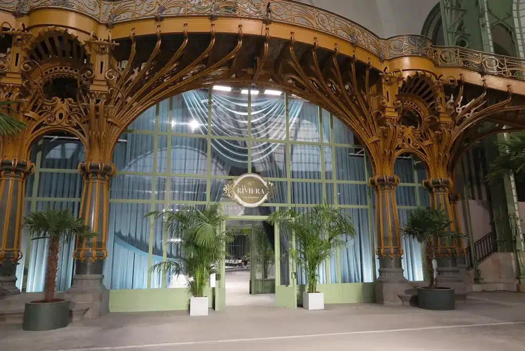 Entrée élégante au Grand Palais, ornée de structures Art nouveau dorées, grands rideaux bleutés et palmiers qui évoquent une ambiance Riviera chic.