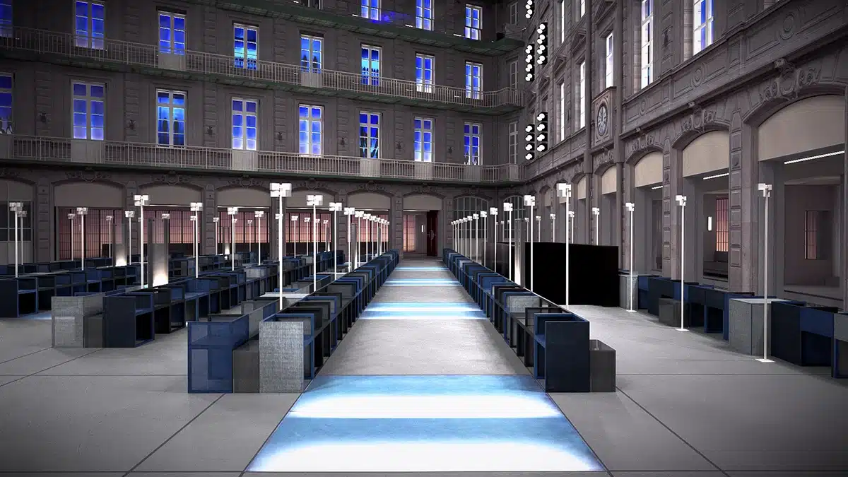 La photo montre un vaste espace intérieur au style architectural classique, transformé en podium contemporain. Une allée centrale illuminée de panneaux lumineux bleutés traverse la pièce, encadrée par des rangées de sièges modernes aux lignes géométriques. Des poteaux élancés équipés de petites lampes rythment le parcours, créant une ambiance de défilé de mode futuriste au cœur d’un bâtiment historique aux hautes fenêtres éclairées d’un bleu vibrant.