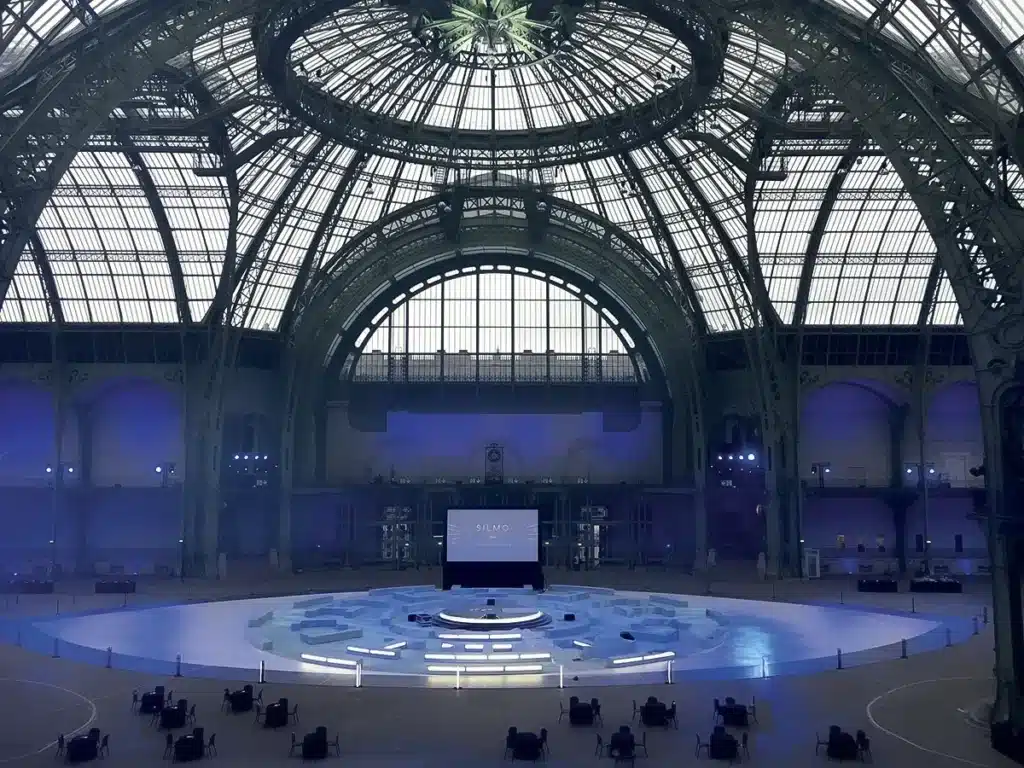 L’image montre une vaste scénographie installée sous la verrière monumentale du Grand Palais, baignée d’une lumière bleutée. Au centre, une grande scène circulaire accueille une structure géométrique composée de plateformes en niveaux, évoquant des blocs de glace ou des formes minérales. Un écran monumental fait face au public, tandis que des éclairages LED soulignent les contours de l’installation. Quelques personnes circulent autour du dispositif, suggérant les préparatifs d’un événement ou d’une présentation. L’ensemble crée une atmosphère contemporaine et spectaculaire, accentuée par l’architecture emblématique du lieu.