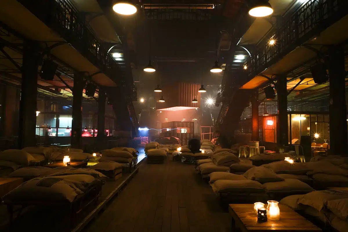 Une atmosphère industrielle immersive, plongée dans une lumière tamisée et chaleureuse. L’espace est rempli de sacs de jute posés sur des palettes, de tables en bois éclairées par des bougies, tandis que les structures métalliques, passerelles et lampes suspendues renforcent l’esthétique d’entrepôt rétro.