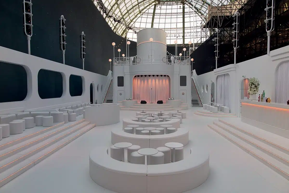 Un décor immaculé inspiré d’un paquebot moderne : grandes courbes blanches, hublots stylisés, gradins arrondis et scène centrale rétro-futuriste baignée d’une lumière douce.