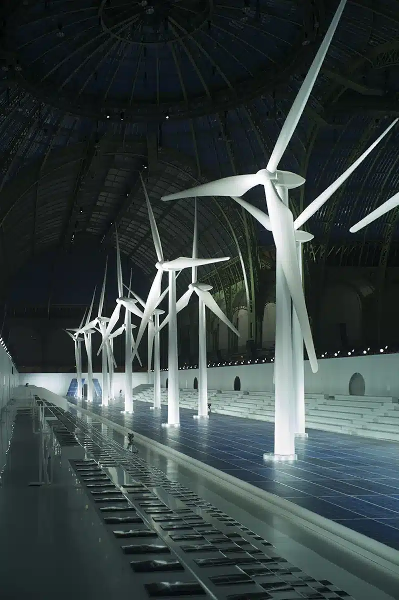 Une immense halle verrière abrite une installation futuriste : une rangée d’éoliennes blanches géantes plantées sur un sol évoquant des panneaux solaires, avec des gradins minimalistes autour. L’ensemble crée un décor spectaculaire autour des énergies renouvelables.