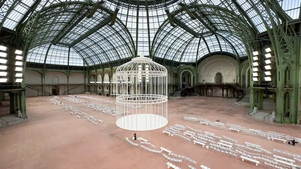 La photo montre une vaste verrière au style monumental, aménagée avec une grande cage blanche au centre, entourée de rangées de chaises et de tables également blanches. L’espace, à la fois majestueux et minimaliste, évoque une scénographie artistique.