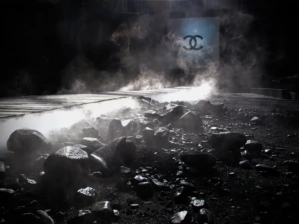Une scénographie sombre et dramatique : un long podium en lames de bois serpente à travers un sol noir parsemé de roches fumantes, créant une atmosphère volcanique. La lumière filtrée sous la verrière du Grand Palais projette des ombres mouvantes sur de hautes tentures grises. Au fond, un écran carré affiche le logo Chanel, accentuant l’aspect mystérieux et cinématographique de la scène.