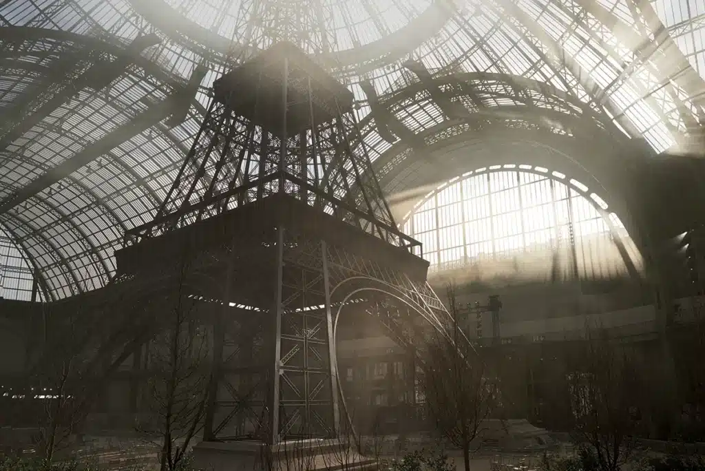 Cette image montre une spectaculaire reproduction monumentale de la tour Eiffel installée sous la nef du Grand Palais. La structure métallique, fidèlement recréée, s’élève vers la verrière qui laisse passer des faisceaux de lumière naturelle, créant une atmosphère dramatique et poétique. La scénographie joue sur l’échelle grandiose de l’iconique monument parisien, immergé dans un environnement légèrement brumeux, avec quelques arbres nus à sa base, comme un jardin urbain réinterprété. L’ensemble compose un décor puissant, où l’architecture du Grand Palais dialogue avec celle de la tour Eiffel dans une mise en scène spectaculaire.