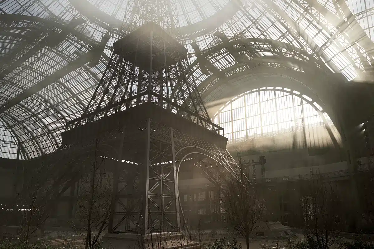 Cette image montre une spectaculaire reproduction monumentale de la tour Eiffel installée sous la nef du Grand Palais. La structure métallique, fidèlement recréée, s’élève vers la verrière qui laisse passer des faisceaux de lumière naturelle, créant une atmosphère dramatique et poétique. La scénographie joue sur l’échelle grandiose de l’iconique monument parisien, immergé dans un environnement légèrement brumeux, avec quelques arbres nus à sa base, comme un jardin urbain réinterprété. L’ensemble compose un décor puissant, où l’architecture du Grand Palais dialogue avec celle de la tour Eiffel dans une mise en scène spectaculaire.