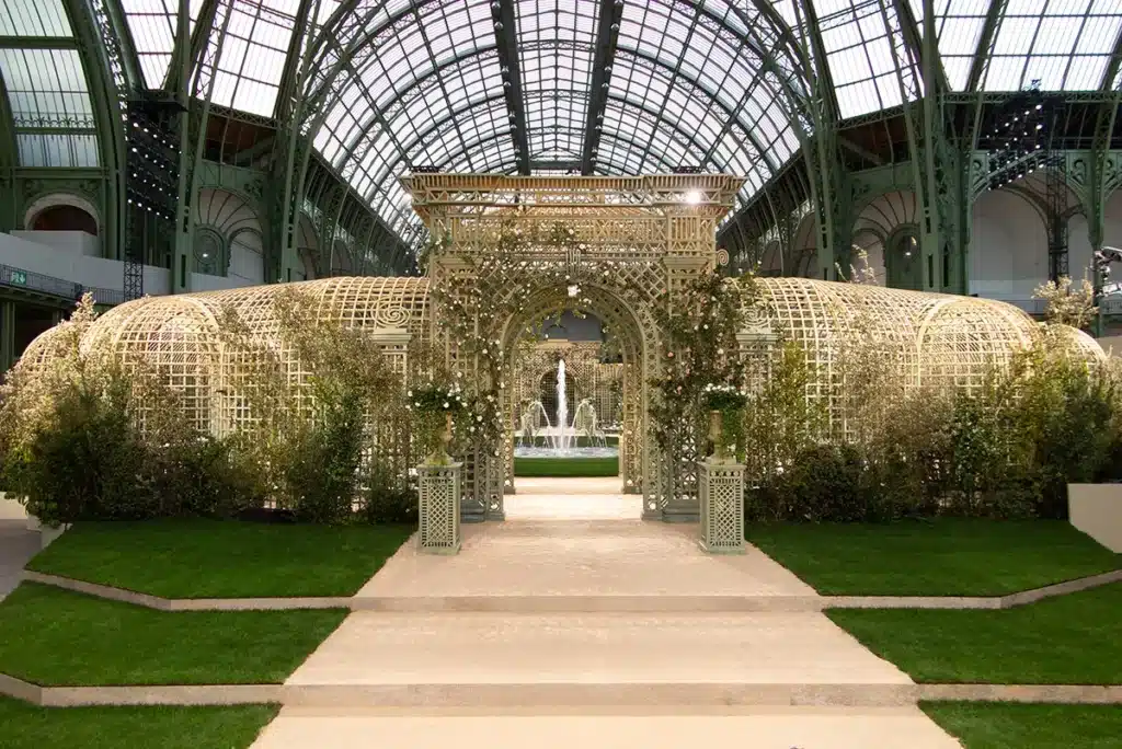 Cette photo montre un décor de défilé transformé en jardin à la française sous la verrière du Grand Palais. Une grande fontaine centrale entourée d’une pelouse parfaitement circulaire structure l’espace, tandis que des treillages ornés de rosiers grimpants forment des galeries élégantes tout autour. Des bancs en arc de cercle accueillent les invités, créant une atmosphère raffinée et romantique inspirée des jardins classiques du XVIIIᵉ siècle.