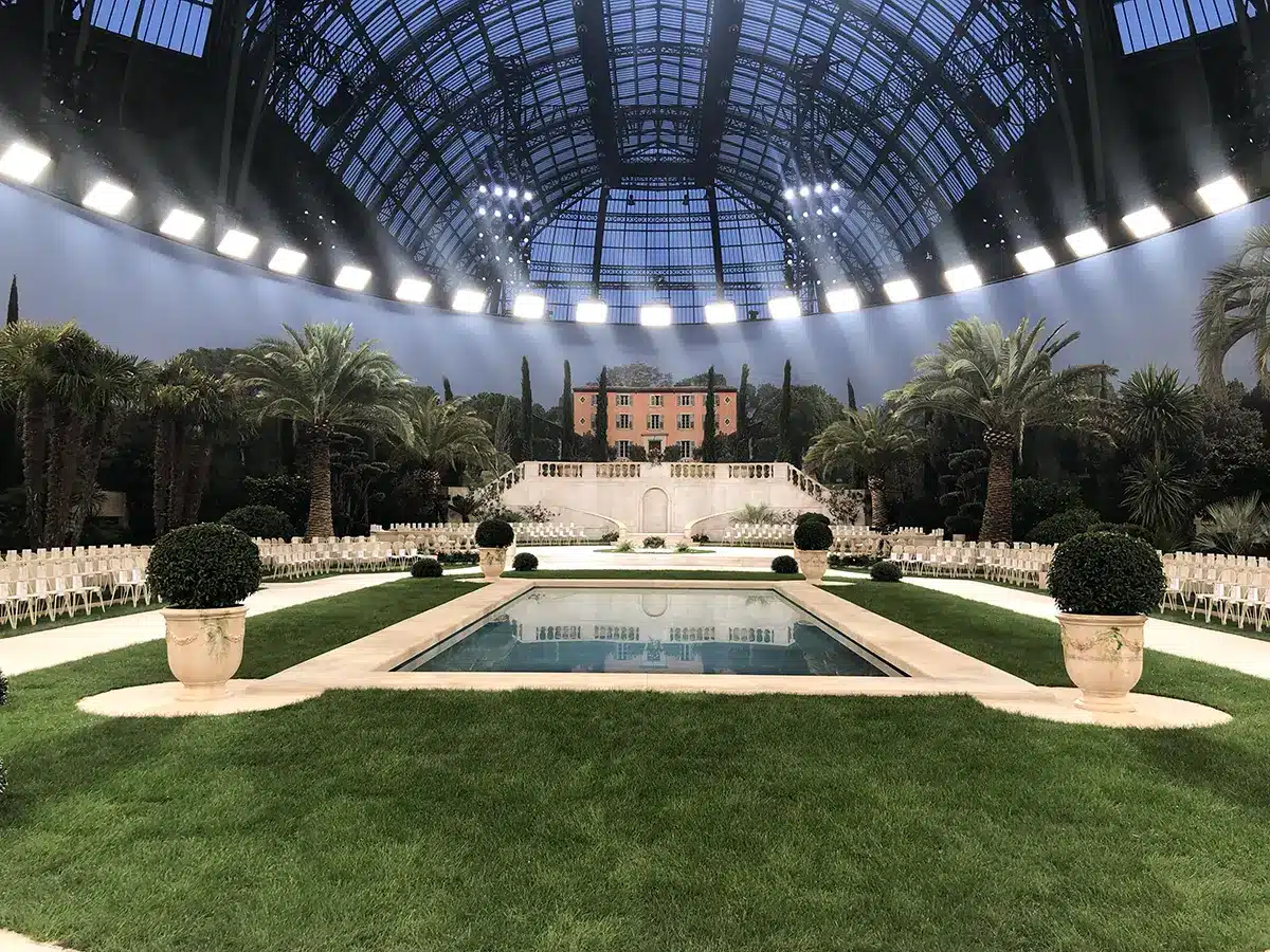 Décor de villa méditerranéenne au Grand Palais, avec piscine centrale, jardins luxuriants, palmiers et ambiance Riviera pour un défilé Chanel.