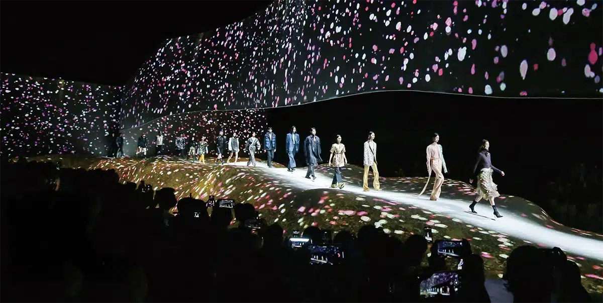 La photo montre un défilé de mode spectaculaire dans un décor immersif. Les mannequins avancent sur un podium ondulé, entièrement recouvert de projections lumineuses évoquant des pétales colorés en chute. Les murs et le plafond sont eux aussi couverts d’images animées, créant un univers visuel enveloppant et poétique. Le public, plongé dans l’obscurité, filme la scène avec ses téléphones, tandis que la passerelle lumineuse met en valeur les silhouettes des modèles. L’ensemble offre une mise en scène futuriste et hautement sensorielle.