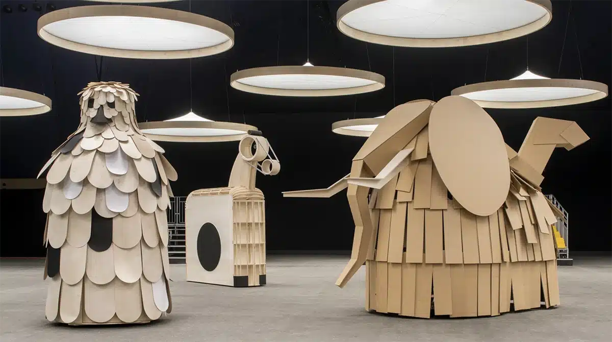 Des sculptures géantes en bois et carton, ressemblant à des animaux stylisés et géométriques, sont exposées dans un espace sombre éclairé par de grands disques lumineux suspendus.