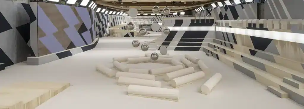 L’image montre un décor de défilé de mode au design très graphique et contemporain. L’espace est habillé de motifs géométriques en camaïeux de gris, beige, noir et lilas, appliqués sur de grandes parois obliques qui encadrent le runway. Au centre, une installation circulaire composée de coussins cylindriques crème disposés en plusieurs anneaux crée une zone sculpturale, presque comme un paysage abstrait. Au-dessus, des sphères métalliques suspendues flottent dans l’air, ajoutant une dimension futuriste et aérienne. Les gradins, réalisés en bois clair et recouverts des mêmes motifs géométriques, s’étirent le long du mur droit. L’ensemble baigne dans une lumière diffuse et uniforme, provenant d’une rangée de projecteurs alignés au plafond, soulignant la pureté des formes et la neutralité des matériaux.