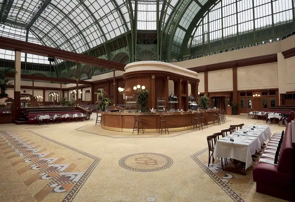 Cette image montre une scénographie monumentale installée sous la verrière du Grand Palais, transformée en brasserie parisienne Belle Époque. Au centre, un vaste bar circulaire en bois et laiton structure l’espace, entouré de banquettes bordeaux, de tables dressées et de mosaïques au sol. L’ensemble évoque une atmosphère chaleureuse, élégante et intemporelle, rappelant l’esthétique raffinée souvent associée aux défilés et décors prestigieux.