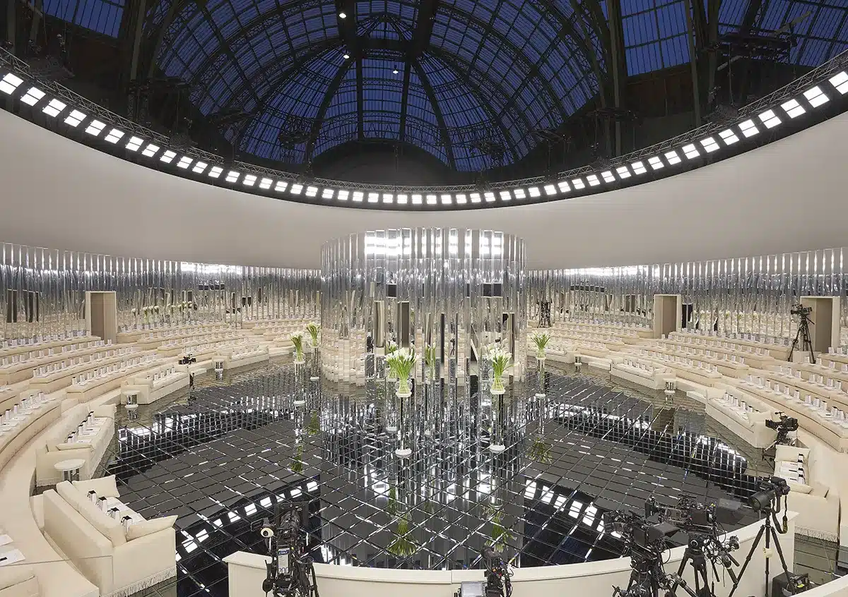 Ce décor présente une mise en scène spectaculaire sous la verrière du Grand Palais : un espace circulaire entièrement enveloppé de miroirs verticaux, créant un jeu infini de reflets. Au centre, une structure cylindrique faite de colonnes miroir capte la lumière et multiplie les perspectives, tandis que le sol en carreaux noirs réfléchissants amplifie l’effet kaléidoscopique. Tout autour, des gradins crème parfaitement symétriques accueillent les invités. L’ensemble compose une atmosphère futuriste, élégante et épurée, où la lumière, le miroir et l’architecture dialoguent pour magnifier la scénographie du défilé haute couture.