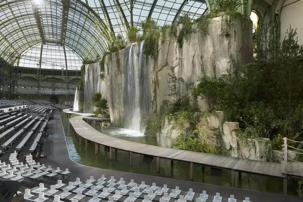 Cette image montre une scénographie spectaculaire installée au Grand Palais, transformé en un immense paysage naturel. Une falaise monumentale en faux rocher occupe tout un côté de l’espace, d’où s’écoulent de grandes cascades d’eau créant un effet immersif et poétique. Au pied de cette paroi, un bassin d’eau accueille un podium en bois courbé, rappelant un chemin naturel serpentant le long de la roche. La végétation — arbres, arbustes, plantes grimpantes — renforce l’impression d’être en pleine nature, contrastant avec la majesté de la verrière métallique du Grand Palais. Des gradins soigneusement alignés entourent le bassin, accueillant les invités du défilé. La lumière naturelle filtre à travers la verrière, illuminant les chutes d’eau et accentuant la dimension spectaculaire du décor. L’ensemble compose une scénographie immersive d’une ampleur exceptionnelle, mêlant nature reconstituée, architecture iconique et mise en scène grandiose.