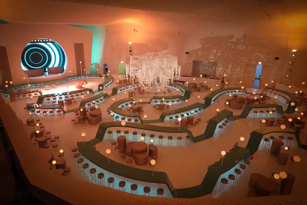 Cette image montre une vaste scénographie immersive conçue comme un lounge futuriste et organique. L’espace, baigné d’une lumière chaude et rosée, est structuré par des parcours sinueux rétroéclairés en bleu, formant des allées fluides où se répartissent des sièges, des tables circulaires et des lampes-boules diffusant une lueur douce. Au centre, un spectaculaire arbre sculptural blanc, semblable à une chute de fleurs suspendues, devient le cœur visuel du décor. En arrière-plan, une scène arrondie et lumineuse encadrée d’un motif circulaire accentue l’esthétique rétro-futuriste. L’ensemble évoque un univers à la fois élégant, onirique et festif, pensé comme un espace de réception spectaculaire.