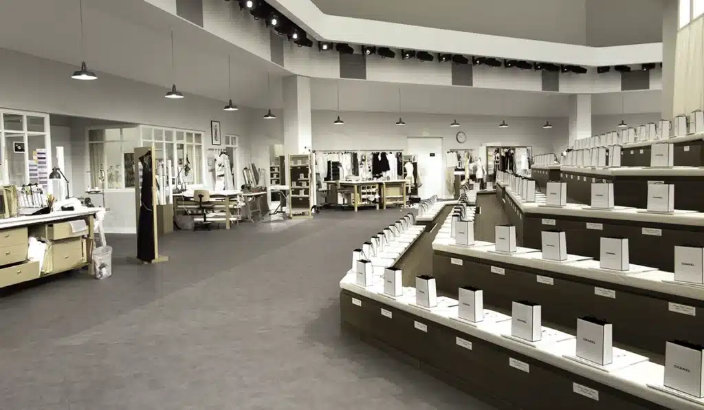 Cette image montre un décor de défilé entièrement recréé comme un atelier de couture Chanel. L’espace est aménagé avec une précision quasi documentaire : grandes tables de travail, bustes de couture, moodboards, portants chargés de prototypes et éclairage industriel diffus par des suspensions métalliques. Sur la droite, les gradins du défilé sont intégrés à la scénographie et disposés comme des postes de travail alignés, chacun équipé de sacs Chanel blancs, évoquant l’idée que les invités prennent place au cœur même du savoir-faire de la Maison. Le décor forme un vaste open-space aux tons neutres — gris, blanc et bois clair — qui met en avant la créativité et la rigueur artisanale du studio, transformant le défilé en une immersion visuelle dans les coulisses de la haute couture.
