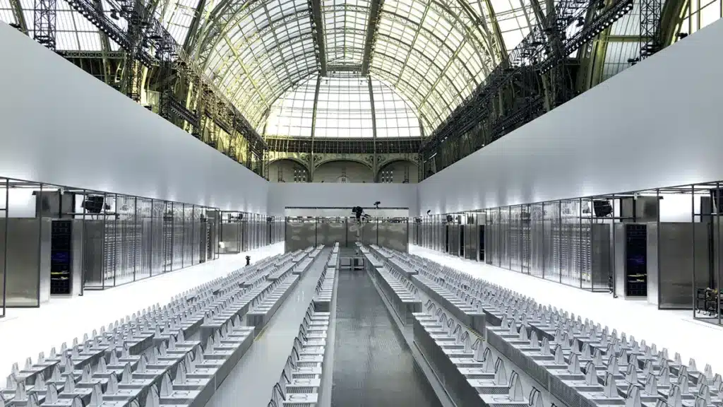 L’image montre un décor de défilé transformé en centre de données futuriste, installé sous la verrière monumentale du Grand Palais. Des rangées impeccables de sièges blancs s’alignent de part et d’autre d’un long podium métallique central, créant une perspective rigoureuse et presque clinique. De chaque côté, des murs composés de baies vitrées renfermant des serveurs et câblages évoquent l’esthétique d’un data center high-tech. Le blanc, l’acier brossé et la lumière froide dominent, accentuant l’atmosphère technologique et ultra-moderne. La structure du Grand Palais, avec ses arches métalliques et sa verrière immense, contraste avec la précision industrielle de l’installation, donnant au décor une présence spectaculaire et quasi cinématographique.