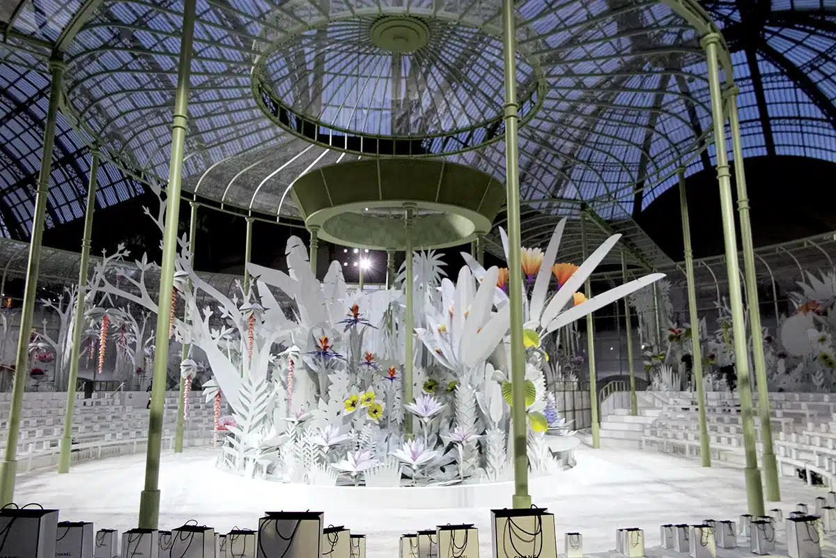 une serre monumentale sous la verrière du Grand Palais, remplie de fleurs géantes en papier et de feuillages découpés. L’univers, poétique et végétal, crée un jardin onirique où la modernité de Chanel rencontre une nature entièrement sculptée et stylisée.