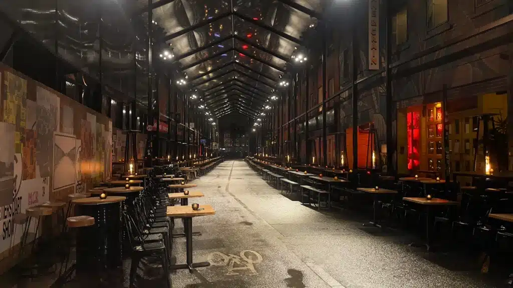 Photo respresentant une rue avec une verrière et des tables et bancs de parts et d'autres.Ambiance de nuit