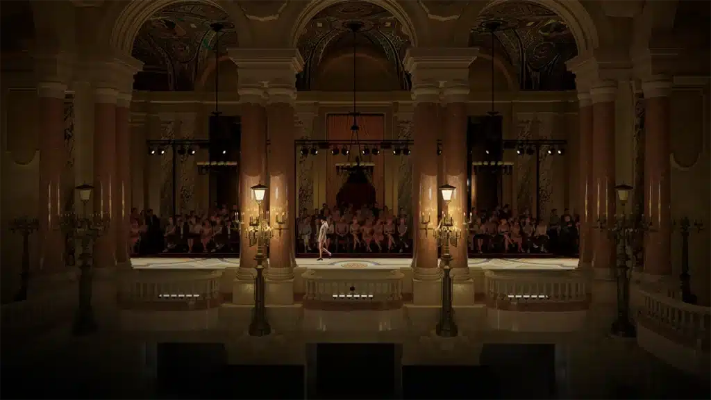 La photo montre un défilé de mode organisé dans un cadre architectural grandiose et historique. De hauts piliers en marbre rose encadrent un podium surélevé où une mannequin marche sous un éclairage théâtral. Derrière les colonnes, le public est assis dans une salle richement décorée de motifs sculptés, de marbres et de plafonds ornés. Des lampadaires anciens et des candélabres dorés ajoutent une touche d’élégance classique, contrastant avec la mise en scène moderne du show. L’ensemble évoque une présentation prestigieuse mêlant luxe, patrimoine et haute couture.