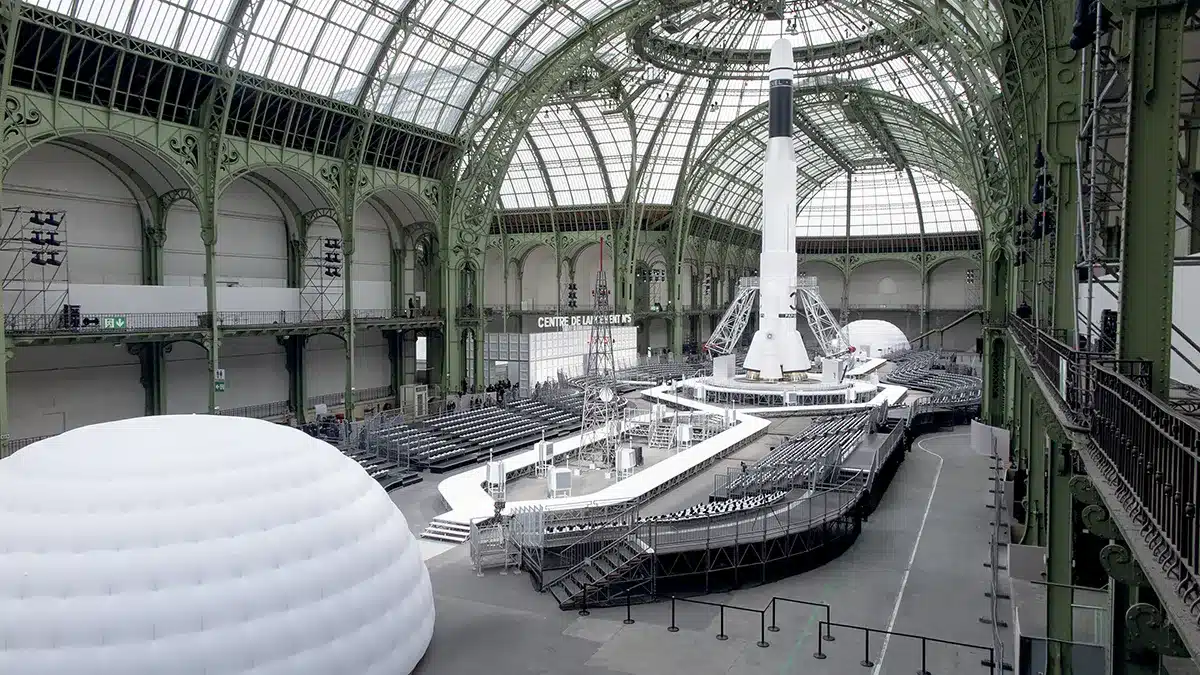 La scénographie représente un pas de tir spatial monumental installé sous la verrière du Grand Palais, avec en son centre une fusée blanche imposante, entourée de passerelles techniques et de gradins circulaires. L’ensemble évoque un centre de lancement futuriste, conçu pour un défilé Chanel imaginé sous la direction de Karl Lagerfeld, mêlant spectacle, innovation et mise en scène grandiose.