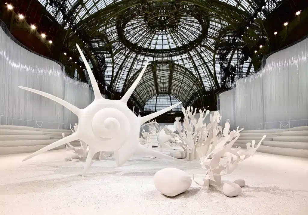 Un décor entièrement blanc transformant le Grand Palais en paysage sous-marin onirique : d’immenses coquillages, coraux stylisés et formes organiques émergent d’un sol clair, baigné par la lumière naturelle filtrant à travers la verrière monumentale. Atmosphère immaculée, douce et irréelle.