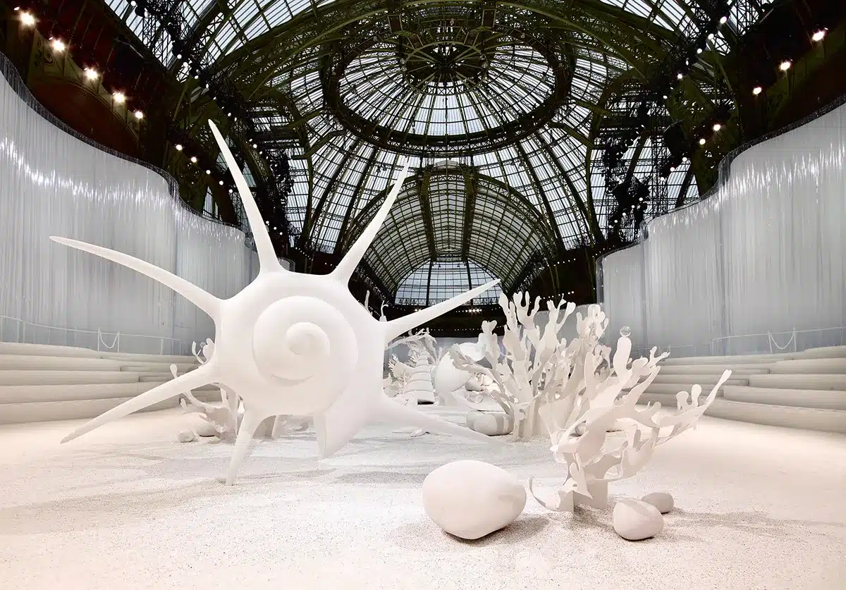 Un décor entièrement blanc transformant le Grand Palais en paysage sous-marin onirique : d’immenses coquillages, coraux stylisés et formes organiques émergent d’un sol clair, baigné par la lumière naturelle filtrant à travers la verrière monumentale. Atmosphère immaculée, douce et irréelle.