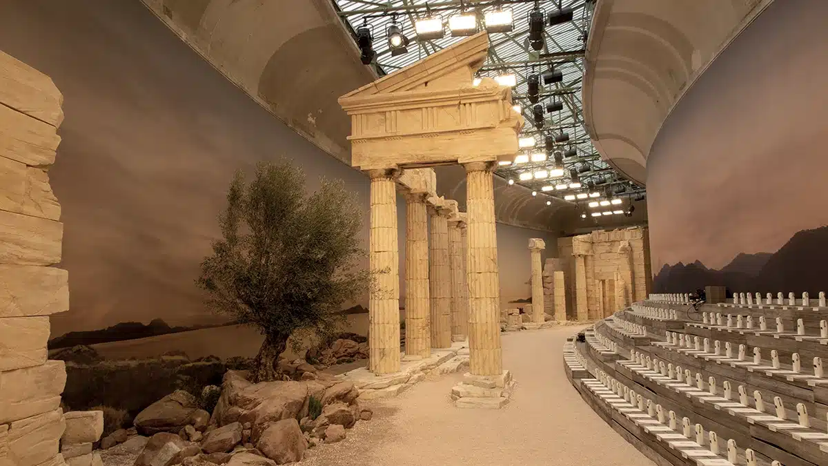 Cette image présente une scénographie monumentale inspirée de la Grèce antique, installée dans un vaste espace sous verrière. On y voit des colonnes en pierre reconstituée, dont un fronton partiellement effondré, évoquant un temple classique. L’allée centrale est recouverte de graviers clairs et ponctuée de rochers et d’un olivier, renforçant l’ambiance méditerranéenne. À droite, des gradins courbés accueillent les spectateurs, tandis qu’un immense fond panoramique peint des montagnes baignées d’une lumière chaude au coucher du soleil. L’ensemble crée un décor architectural puissant et élégant, rappelant les paysages et les ruines antiques de la Grèce.