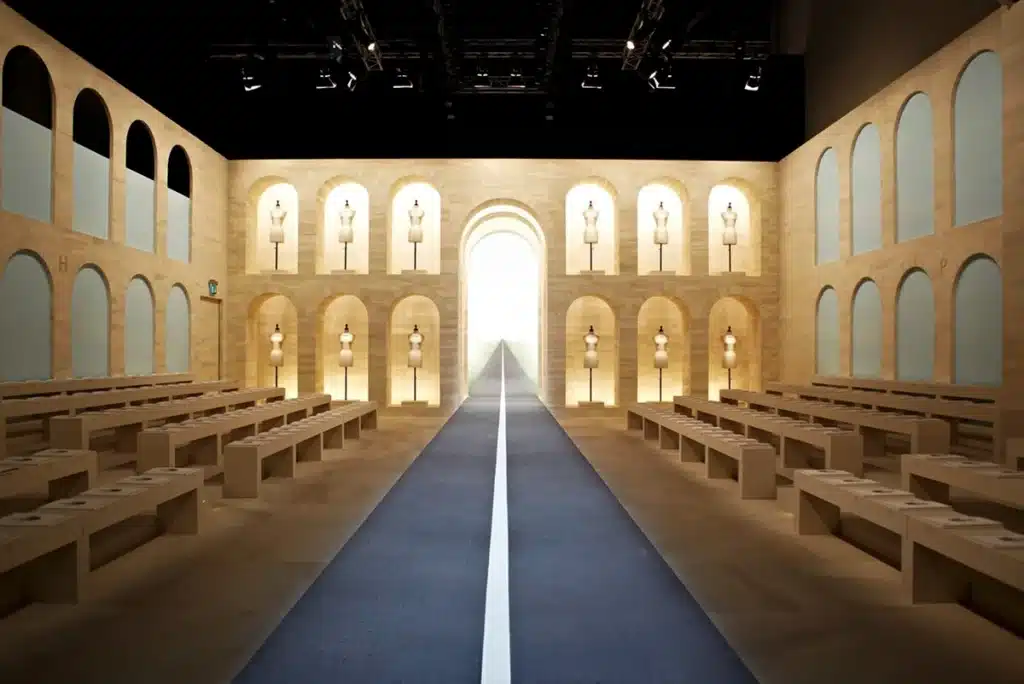 un long podium encadré de murs beiges à arches répétées, chacune abritant un buste de couture éclairé. L’ensemble crée une ambiance architecturale, symétrique et très couture, comme un cloître moderne dédié à l’art du vêtement.