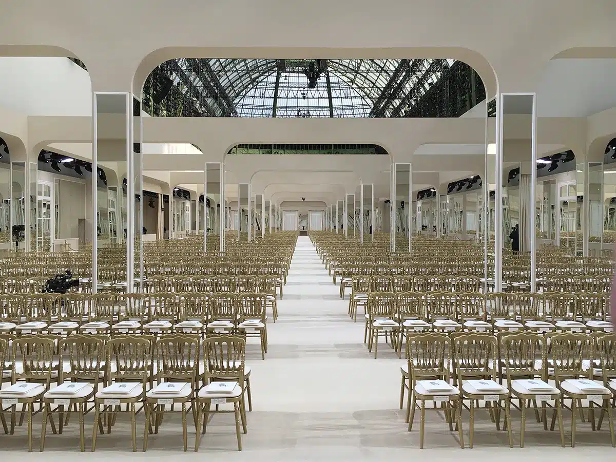 Cette image montre un décor de défilé installé sous la verrière du Grand Palais, composé de rangées impeccablement alignées de chaises dorées formant un vaste espace symétrique. Au centre, un long podium blanc s’étend vers le fond, encadré par de hautes colonnes miroir qui reflètent la lumière et l’architecture du lieu. L’ensemble crée une atmosphère élégante et grandiose, mêlant modernité épurée et classicisme du cadre historique.