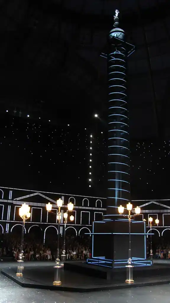 une immense reconstitution lumineuse de la colonne Vendôme, installée au centre du Grand Palais. Elle est éclairée par des lignes LED blanches, qui soulignent sa spirale et son socle. Autour, des réverbères transparents diffusent une lumière dorée. Le fond noir parsemé de points lumineux façon ciel étoilé renforce l’effet nocturne parisien. Les façades de la place, dessinées en lignes lumineuses blanches, encadrent la scène et donnent l’illusion d’un Paris stylisé et futuriste.