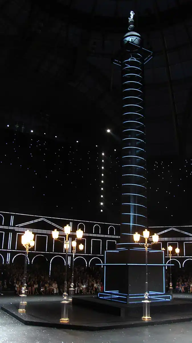 une immense reconstitution lumineuse de la colonne Vendôme, installée au centre du Grand Palais. Elle est éclairée par des lignes LED blanches, qui soulignent sa spirale et son socle. Autour, des réverbères transparents diffusent une lumière dorée. Le fond noir parsemé de points lumineux façon ciel étoilé renforce l’effet nocturne parisien. Les façades de la place, dessinées en lignes lumineuses blanches, encadrent la scène et donnent l’illusion d’un Paris stylisé et futuriste.