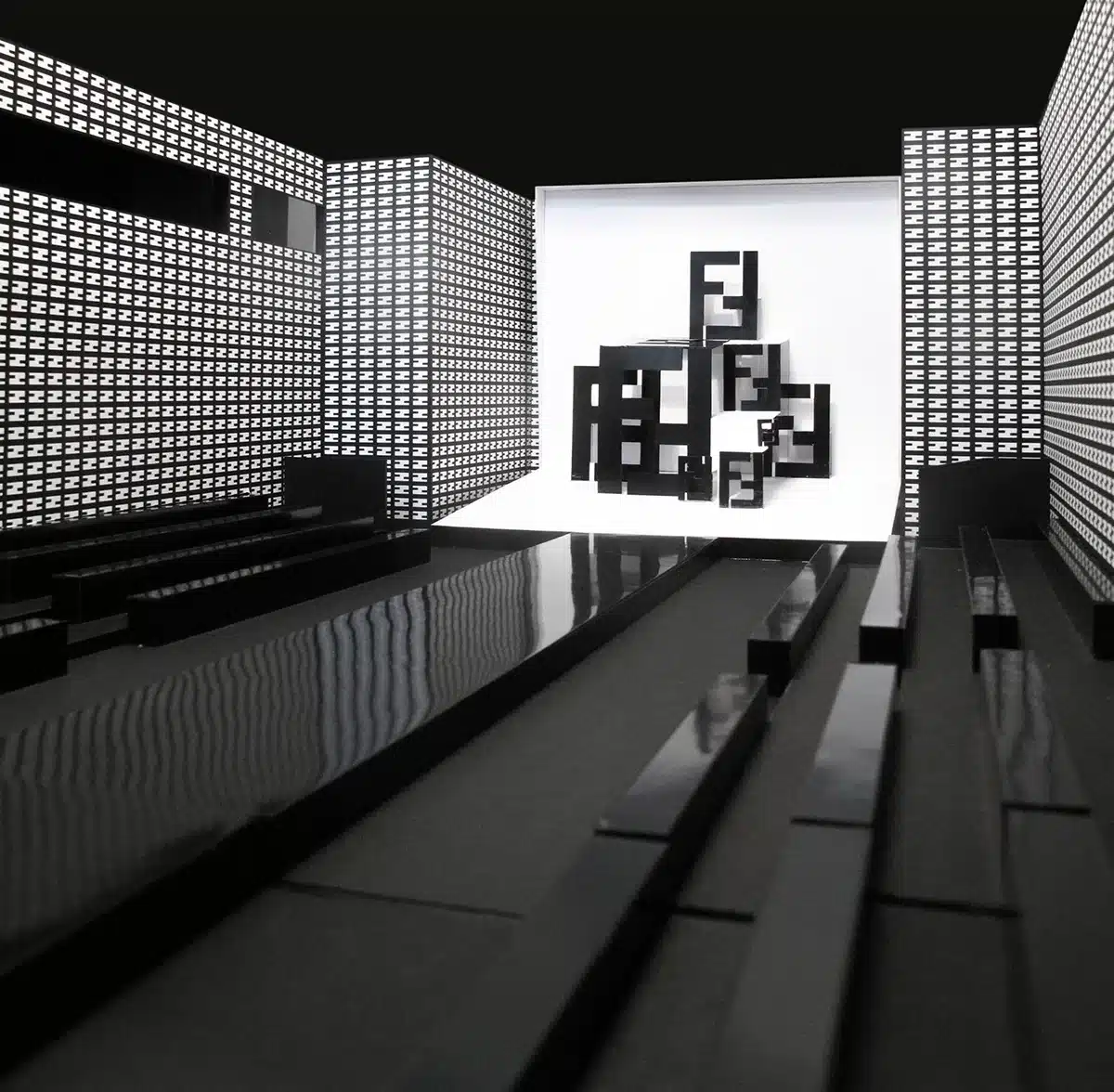 Un décor de défilé Fendi ultra-graphique et minimaliste : un long podium noir brillant encadré de murs couverts du motif FF répété, menant à une sculpture centrale de logos FF imbriqués en noir et blanc. L’ensemble crée une ambiance futuriste, géométrique et très identitaire.