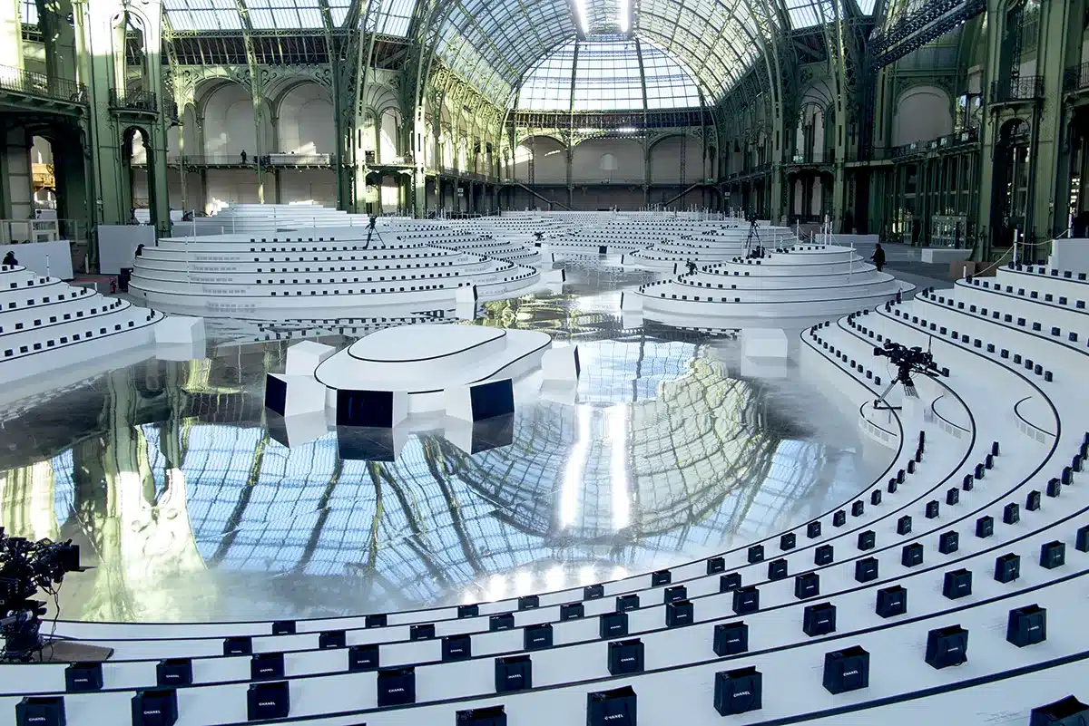 La photo montre une scénographie spectaculaire installée sous la verrière du Grand Palais. L’espace est aménagé en vastes gradins circulaires blancs, disposés en anneaux concentriques autour d’une scène centrale aux formes organiques. Le sol, entièrement recouvert d’une surface miroir, reflète l’architecture monumentale du lieu et crée un effet visuel saisissant. Sur chaque siège repose un sac noir siglé Chanel, soulignant le caractère exclusif de l’événement. Des caméras sont disposées çà et là, prêtes à capturer le défilé ou la présentation à venir. L’ensemble évoque un univers futuriste, épuré et d’une grande élégance.