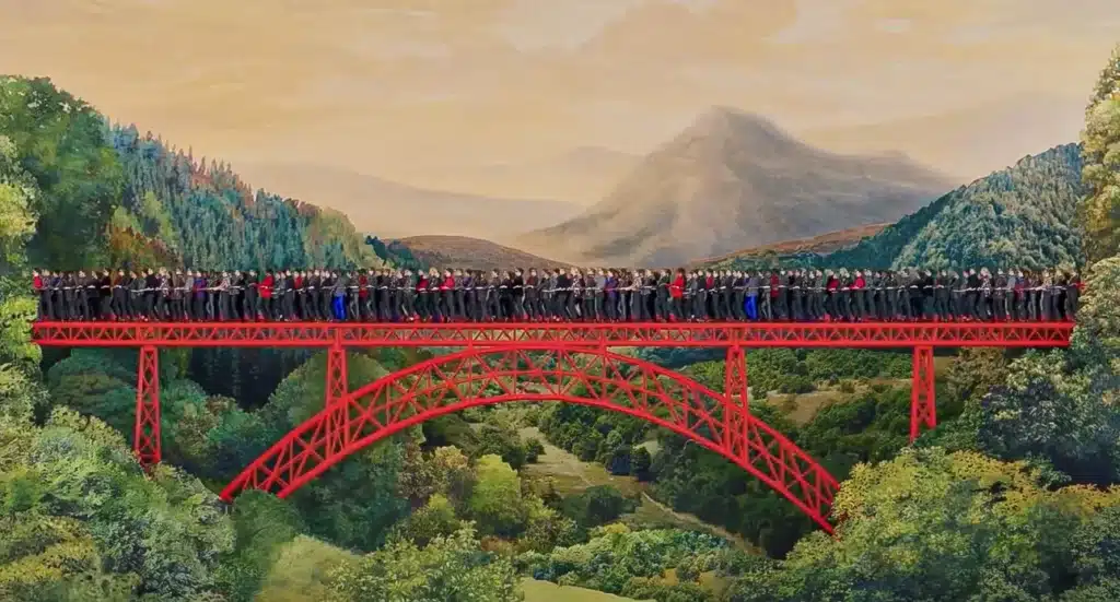 La photo montre un long pont métallique rouge traversant une vallée verdoyante entourée de collines et de montagnes au loin. Le pont est entièrement rempli d’un grand groupe de personnes alignées côte à côte, créant une impression de foule dense et continue. Le paysage, baigné d’une lumière douce, contraste avec la structure vive du pont, offrant une scène à la fois spectaculaire et harmonieuse entre architecture et nature.