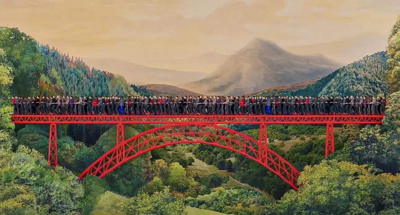 La photo montre un long pont métallique rouge traversant une vallée verdoyante entourée de collines et de montagnes au loin. Le pont est entièrement rempli d’un grand groupe de personnes alignées côte à côte, créant une impression de foule dense et continue. Le paysage, baigné d’une lumière douce, contraste avec la structure vive du pont, offrant une scène à la fois spectaculaire et harmonieuse entre architecture et nature.