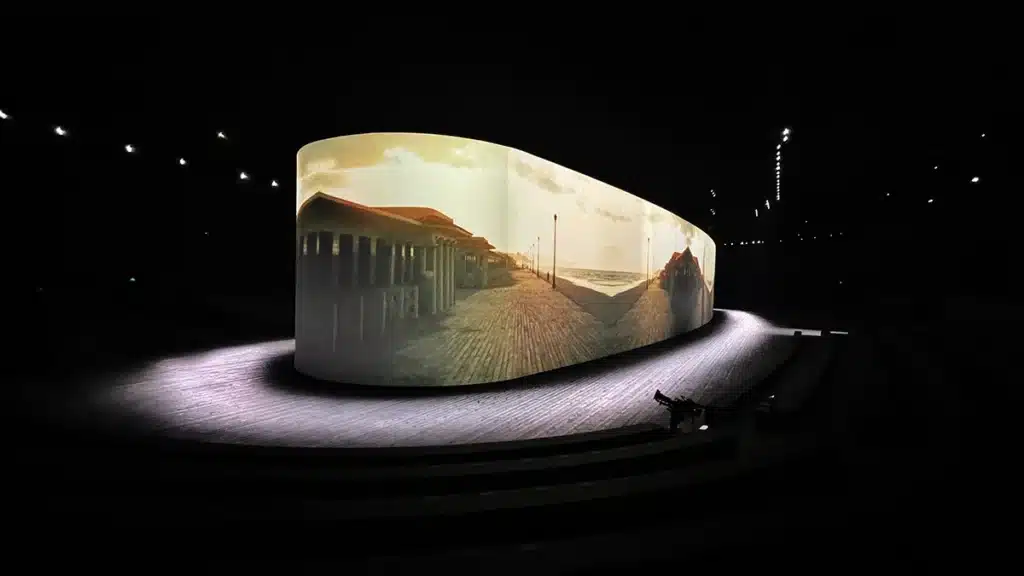 La photo montre un vaste espace sombre où trône un immense écran panoramique courbé, légèrement ovale, placé au centre d’une scène circulaire. Sur cette surface monumentale est projetée une vidéo immersive montrant une promenade en bois menant vers la mer, baignée d’une lumière dorée de coucher de soleil, avec des silhouettes en surimpression. Des gradins minimalistes encerclent partiellement la zone, tandis que des projecteurs ponctuent le plafond d’une ligne lumineuse. L’ensemble évoque une installation artistique ou une scénographie immersive au style cinématographique.