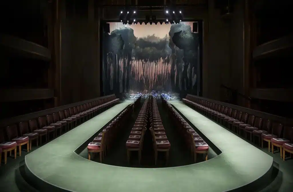 Podium en U dans un théâtre sombre, éclairage dramatique et décor peint évoquant une forêt mystérieuse en arrière-plan.