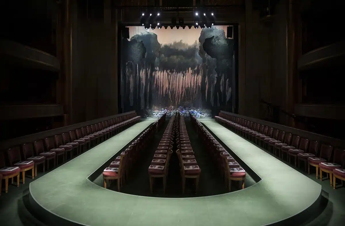 Podium en U dans un théâtre sombre, éclairage dramatique et décor peint évoquant une forêt mystérieuse en arrière-plan.