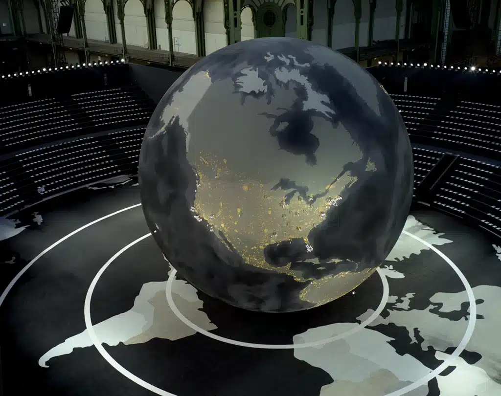 Un immense globe terrestre occupe le centre de la scène, présenté comme une sphère monumentale sombre où brillent des milliers de lumières évoquant les villes du monde la nuit. Il repose sur un sol cartographié représentant les continents en tons gris. Tout autour, des gradins circulaires accueillent les spectateurs. L’ensemble crée une atmosphère spectaculaire et immersive, évoquant à la fois la planète, la mondialisation et une vision futuriste du monde.