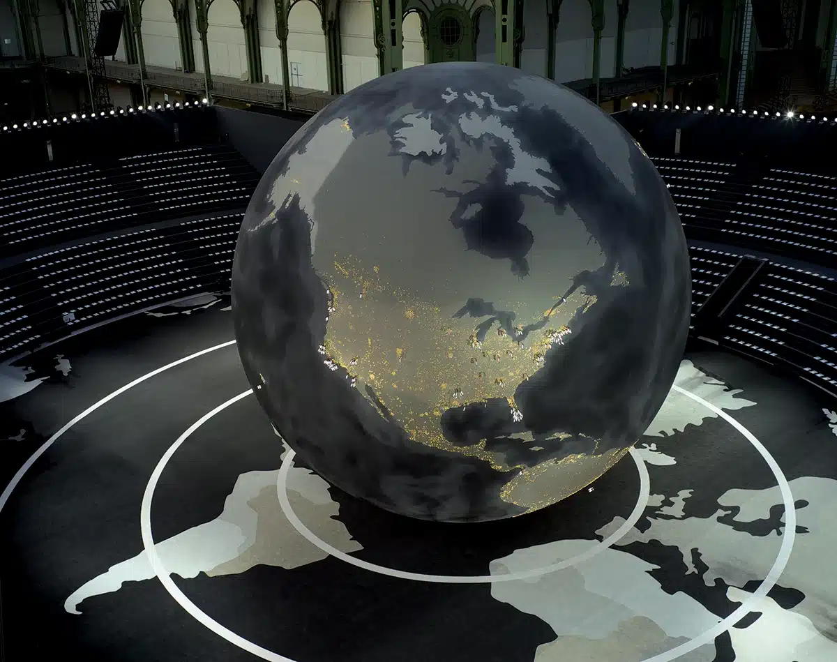 Un immense globe terrestre occupe le centre de la scène, présenté comme une sphère monumentale sombre où brillent des milliers de lumières évoquant les villes du monde la nuit. Il repose sur un sol cartographié représentant les continents en tons gris. Tout autour, des gradins circulaires accueillent les spectateurs. L’ensemble crée une atmosphère spectaculaire et immersive, évoquant à la fois la planète, la mondialisation et une vision futuriste du monde.