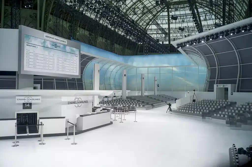 La scénographie représente un hall d’aéroport entièrement réinventé sous la verrière du Grand Palais. L’espace, lumineux et immaculé, est transformé en terminal Chanel Airlines, avec comptoirs d’enregistrement numérotés, portiques, rubans de guidage et un grand panneau d’affichage des départs portant la mention « Aéroport Paris Cambon ». Les parois courbes, traitées comme d’immenses baies vitrées ouvertes sur un ciel bleu artificiel, amplifient l’illusion d’un véritable terminal moderne. Les gradins, parfaitement alignés, accueillent les invités comme des passagers en attente d’embarquement. L’ensemble combine esthétique minimaliste, codes du voyage et précision architecturale, créant un décor immersif et futuriste typique des défilés Chanel orchestrés avec un sens aigu du détail.