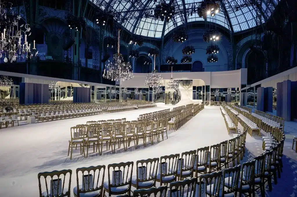 Cette image dévoile le somptueux décor d’un défilé Chanel installé sous la verrière monumentale du Grand Palais. La scénographie, à la fois élégante et théâtrale, transforme l’espace en un univers lumineux et raffiné. Au centre, un podium immaculé se déploie en courbe, menant vers un escalier monumental recouvert de blanc, encadré par des panneaux de miroirs qui reflètent la lumière et agrandissent visuellement le décor. Des lustres en cristal, suspendus en hauteur tels des bijoux scintillants, apportent une atmosphère fastueuse et classique. Les rangées de chaises dorées parfaitement alignées forment un cadre harmonieux et sophistiqué ; chacune est équipée d’un petit sac signé Chanel, réservé aux invités. Le sol clair accentue la sensation d’espace et de pureté. L’ensemble respire le luxe français, combinant architecture grandiose, éclairage théâtral et codes intemporels de Chanel pour accueillir un défilé d’exception dans un écrin spectaculaire.