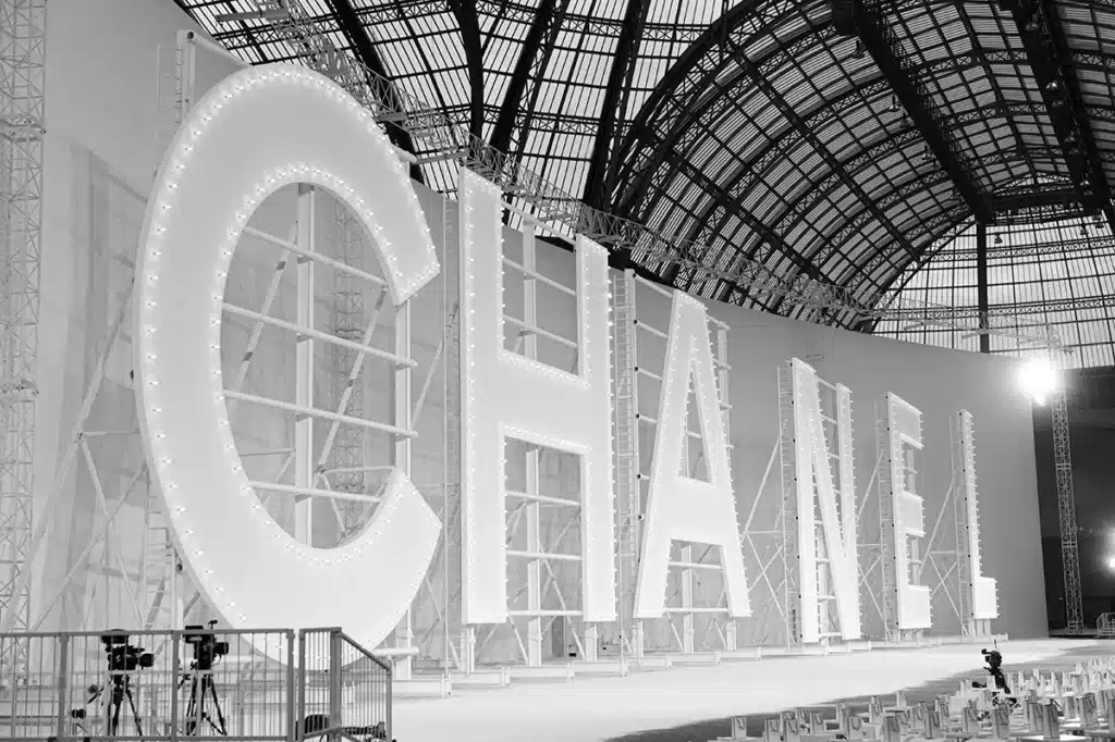 La photo montre l’intérieur monumental du Grand Palais, où d’immenses lettres blanches formant le mot CHANEL sont installées comme un décor spectaculaire. Chaque lettre, rétroéclairée par une multitude de petites ampoules, évoque les enseignes lumineuses hollywoodiennes. La structure métallique qui les soutient contraste avec la verrière majestueuse du bâtiment. Au sol, des rangées de sièges encore vides et plusieurs caméras suggèrent la préparation d’un défilé de mode de grande envergure. L’ensemble, en noir et blanc, renforce l’élégance et l’impact visuel de la scène.